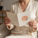 Femme vêtue d'un ensemble crème satiné tenant une carte de vœux ouverte, photographiée sans visage visible. La carte montre un cercle rose poudré aux lignes concentriques avec une feuille verte, sur fond intérieur chaleureux aux tons neutres.