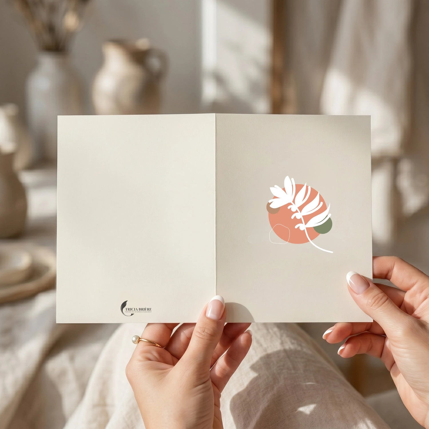 Deux mains féminines aux ongles naturels tenant une carte de vœux ouverte devant un décor intérieur flou aux tons beige et crème. La carte présente une illustration botanique abstraite en terracotta, blanc et vert sauge sur fond ivoire.