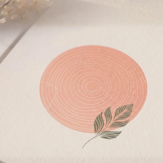 Gros plan sur la couverture d'une carte de vœux posée en angle sur une surface texturée ivoire. L'illustration centrale est un grand cercle rose poudré avec des cernes concentriques blancs et une feuille tropicale vert sauge, avec des fleurs séchées blanches en arrière-plan.