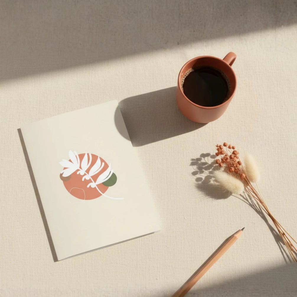 Vue aérienne d'une carte de vœux ouverte sur une surface en lin naturel, accompagnée d'une tasse de café terracotta, d'un crayon en bois et de fleurs séchées beige. La carte présente une illustration botanique abstraite en terracotta et vert sauge.