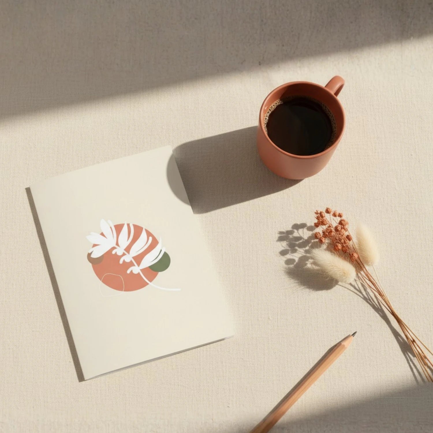 Vue aérienne d'une carte de vœux ouverte sur une surface en lin naturel, accompagnée d'une tasse de café terracotta, d'un crayon en bois et de fleurs séchées beige. La carte présente une illustration botanique abstraite en terracotta et vert sauge.