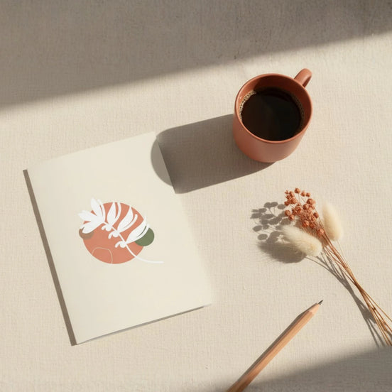 Vue aérienne d'une carte de vœux ouverte sur une surface en lin naturel, accompagnée d'une tasse de café terracotta, d'un crayon en bois et de fleurs séchées beige. La carte présente une illustration botanique abstraite en terracotta et vert sauge.