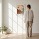 Femme de dos contemplant l'affiche artistique « Écho & Métamorphose » par Tricia Brière dans un cadre blanc, accrochée sur un mur baigné de lumière naturelle.