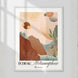 Affiche « Écho & Métamorphose » par Tricia Brière dans un cadre en bois blanc. Paysage abstrait avec montagnes terracotta, feuillage vert et sphères célestes sur fond pêche.