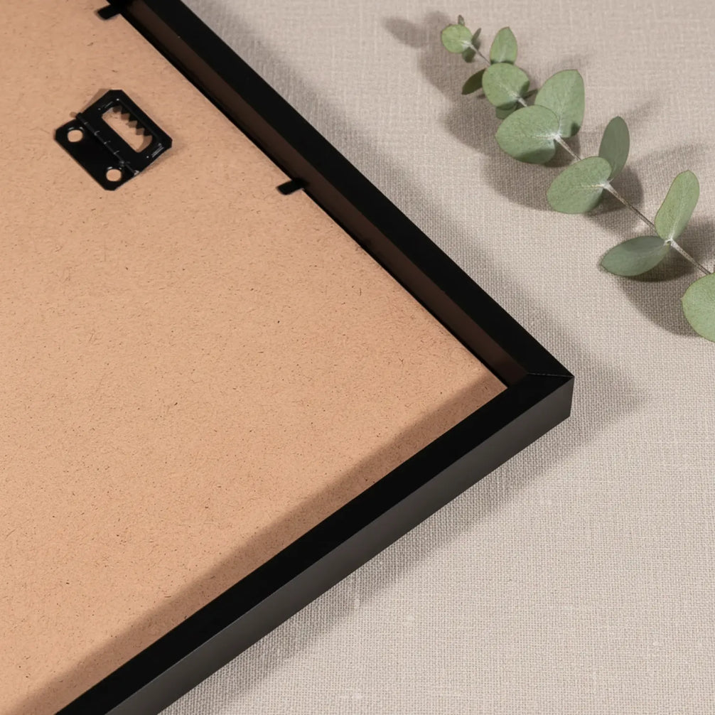 Gros plan sur le dos d'un cadre en bois noir montrant le système d'accroche métallique intégré, posé sur une surface en lin beige avec une branche d'eucalyptus en arrière-plan.