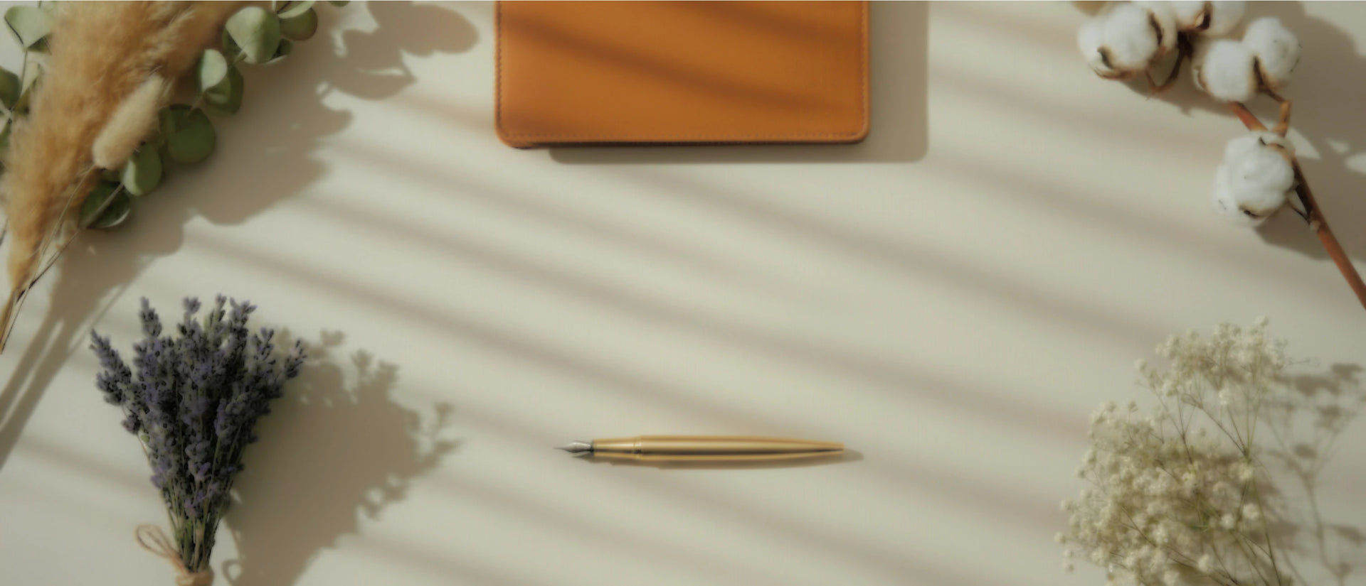 Vue de dessus (flat lay) d'un espace de travail minimaliste et chaleureux sur fond beige, baigné par des ombres d'un store vénitien. Au centre se trouve un stylo-plume doré, surmonté d'un carnet en cuir marron. Le cadre est entouré d'éléments naturels : un bouquet de lavande séchée en bas à gauche, de l'eucalyptus en haut à gauche, une branche de coton en haut à droite, et du gypsophile blanc en bas à droite.