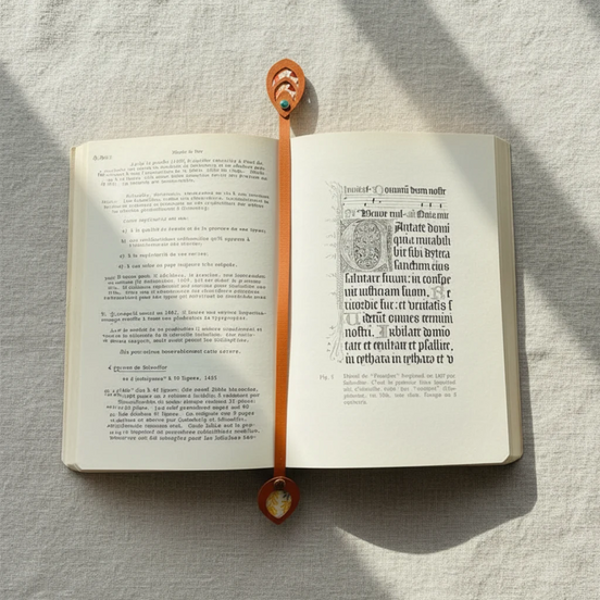 Marque-page artisanal en cuir cognac traversant verticalement un livre ancien ouvert sur une enluminure gothique, son médaillon supérieur ovale à découpes concentriques avec rivet vert émeraude visible en haut de la page et son médaillon inférieur en forme de goutte avec papier japonais fleuri et rivet ambré dépassant en bas, en lumière matinale rasante sur fond lin blanc.