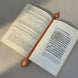 Marque-page artisanal en cuir cognac posé à plat dans un livre ancien ouvert, sa lanière s'étirant sur toute la largeur des pages avec une tête losange incrustée de papier japonais en haut et un médaillon ovale fleuri avec rivet ambré en bas, en lumière naturelle douce.