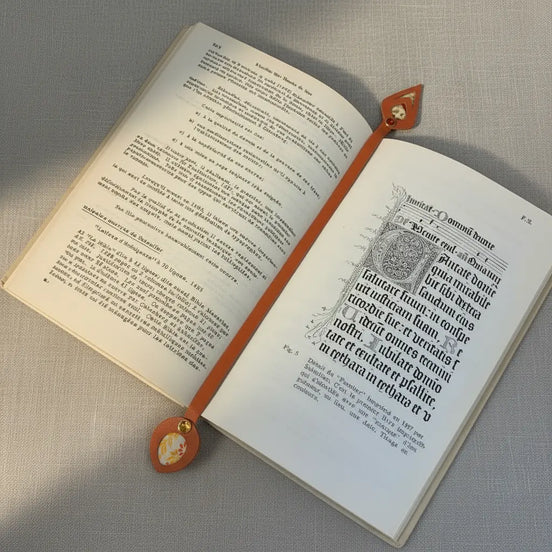 Marque-page artisanal en cuir cognac posé à plat dans un livre ancien ouvert, sa lanière s'étirant sur toute la largeur des pages avec une tête losange incrustée de papier japonais en haut et un médaillon ovale fleuri avec rivet ambré en bas, en lumière naturelle douce.
