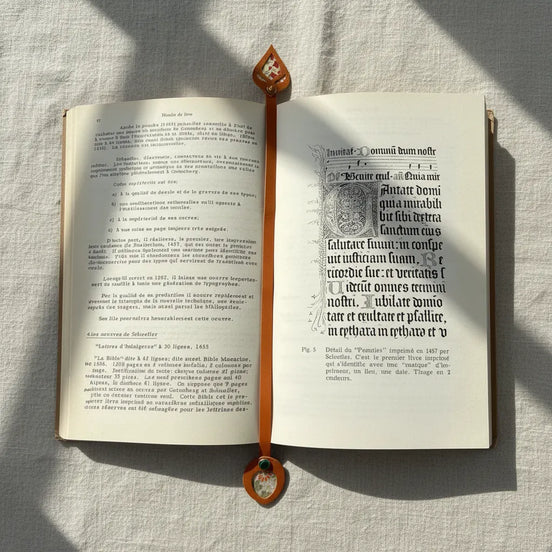 Marque-page en cuir cognac posé dans un livre ancien ouvert, avec médaillon à découpe feuille et rivet au sommet et en bas de la lanière, entouré de fleurs séchées et branches d'eucalyptus en lumière naturelle.