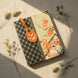 Marque-page en cuir cognac glissé dans un carnet à couverture japonaise noire et florale rouge-orange, son médaillon ovale en haut avec rivet vert émeraude et papier japonais fleuri orange-crème visible, en lumière solaire dorée sur fond lin.