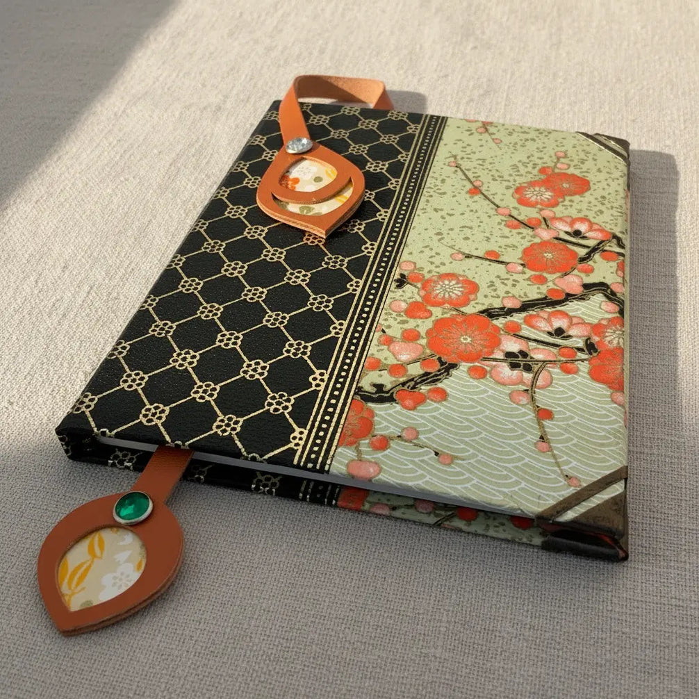 Marque-page artisanal en cuir cognac posé en diagonale sur un livre ancien ouvert, avec un médaillon ovale incrusté de tissu japonais fleuri jaune-orange et un rivet ambré en bas, et une tête en forme de losange avec rivet rouge en haut, en lumière matinale rasante.
