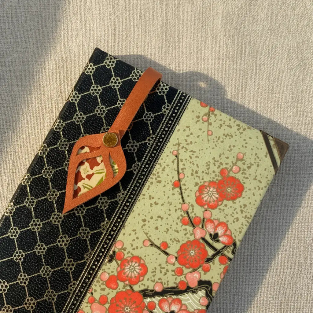 Marque-page artisanal en cuir cognac découpé en forme de feuille, posé sur un carnet à couverture japonaise noire et dorée avec tissu fleuri rouge.