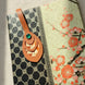 Gros plan sur le médaillon supérieur d'un marque-page artisanal en cuir cognac posé sur un carnet à couverture japonaise noire et florale rouge : médaillon ovale à trois niveaux découpés révélant un papier japonais fleuri orange-crème, orné d'un rivet vert émeraude, en lumière solaire rasante.