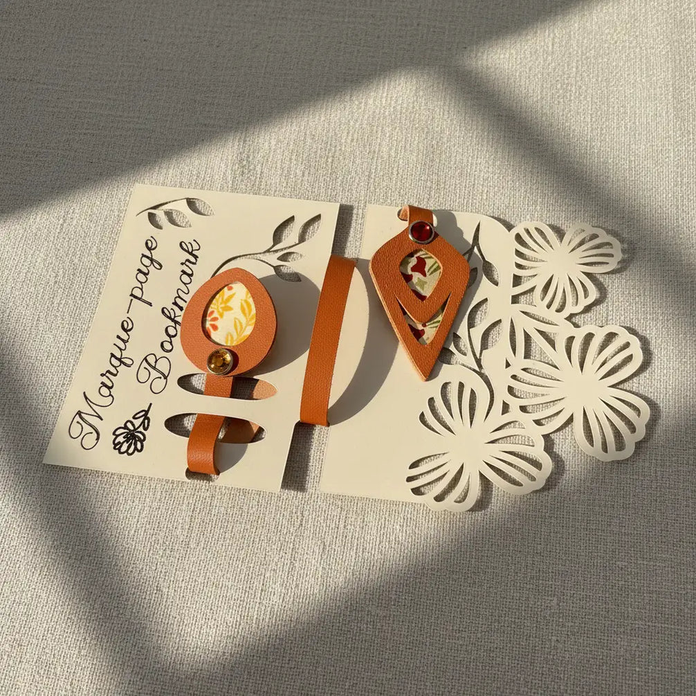Marque-pages artisanal en cuir cognac présentés côte à côte sur leur carte d'emballage découpée : l'un à médaillon ovale avec papier japonais fleuri jaune-vert et rivet vert émeraude, l'autre à tête losange avec papier floral et rivet rouge rubis, ombres géométriques douces sur fond lin.