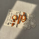 Marque-page artisanal en cuir cognac sur carte d'emballage découpée floralement : médaillons à découpe feuille et ovale incrustés de papier japonais à motifs fleuris jaune et orange, ornés de rivets vert et cristal.