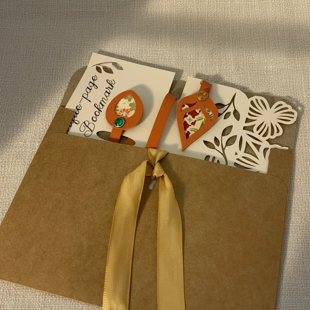 Marque-page en cuir cognac en forme de feuille découpée, présentée dans une enveloppe kraft fermée d'un ruban doré, avec une carte illustrée de motifs floraux.