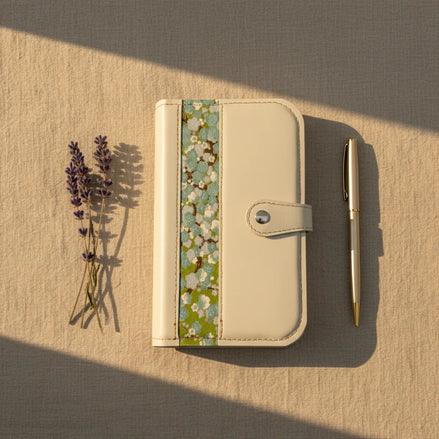Prise de vue aérienne d'un organisateur modulaire fermé en cuir blanc cassé, disposé verticalement sur un textile de fond beige. À sa droite sont alignés un stylo fin en métal doré et de délicates tiges de lavandes séchées. Une lumière naturelle rasante baigne la scène de gauche à droite, projetant des ombres douces pour une atmosphère calme et sereine.