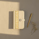 Photographie en plongée sur un fond texturé beige d'un organisateur modulaire en cuir écru fermé, orné d'une bande centrale à motifs floraux et végétaux. Un stylo doré et un bouquet séché de gypsophile sont disposés sur la droite, le tout baigné d'une lumière douce.