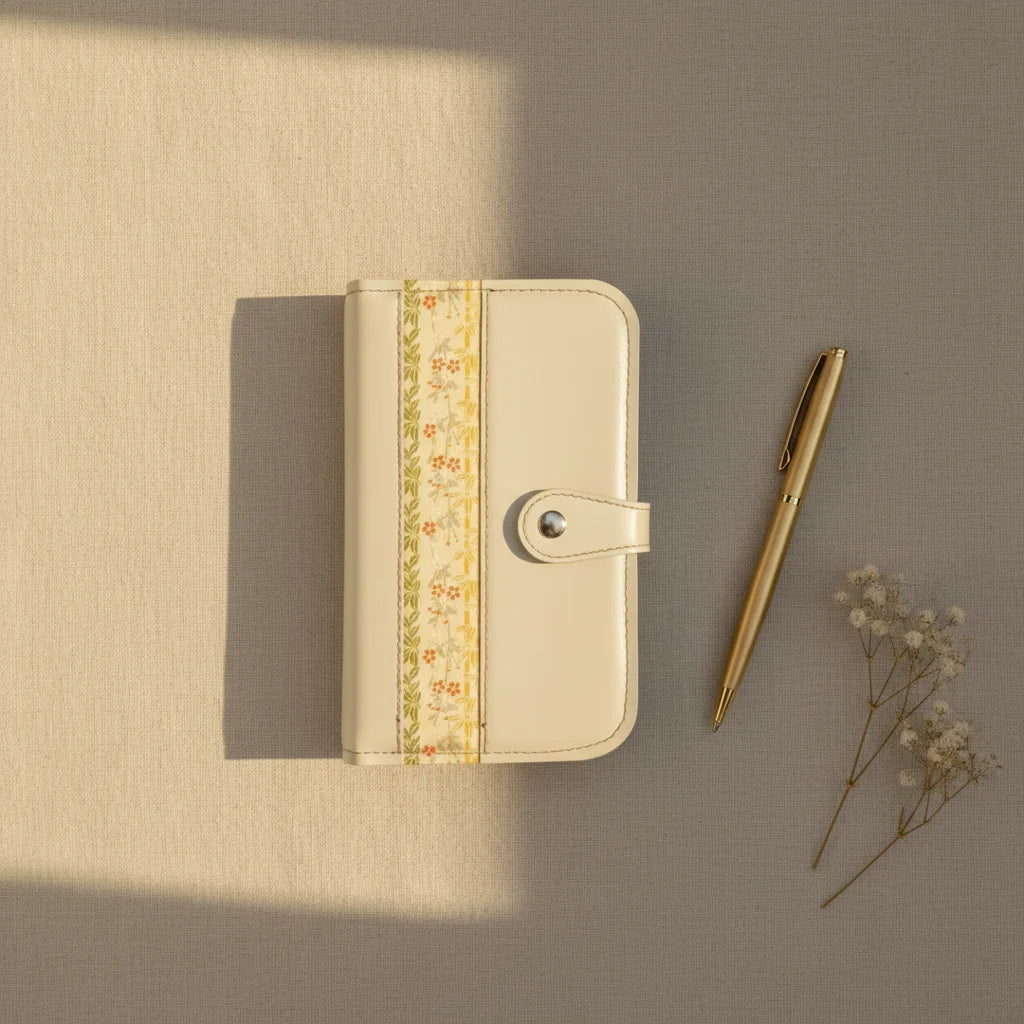 Photographie en plongée sur un fond texturé beige d'un organisateur modulaire en cuir écru fermé, orné d'une bande centrale à motifs floraux et végétaux. Un stylo doré et un bouquet séché de gypsophile sont disposés sur la droite, le tout baigné d'une lumière douce.