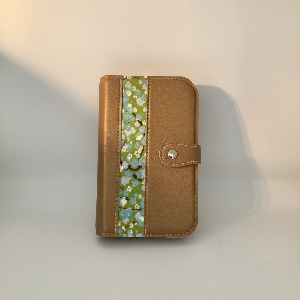 Une photo professionnelle en gros plan d'un classeur modulaire fermé, de couleur marron doré avec une texture grainée. Une bande centrale en tissu présente un motif délicat de fleurs de cerisier bleu ciel et blanches sur un fond vert vif. Le fermoir à pression argenté est clairement visible sur le côté droit. L'objet est posé sur un fond gris neutre.
