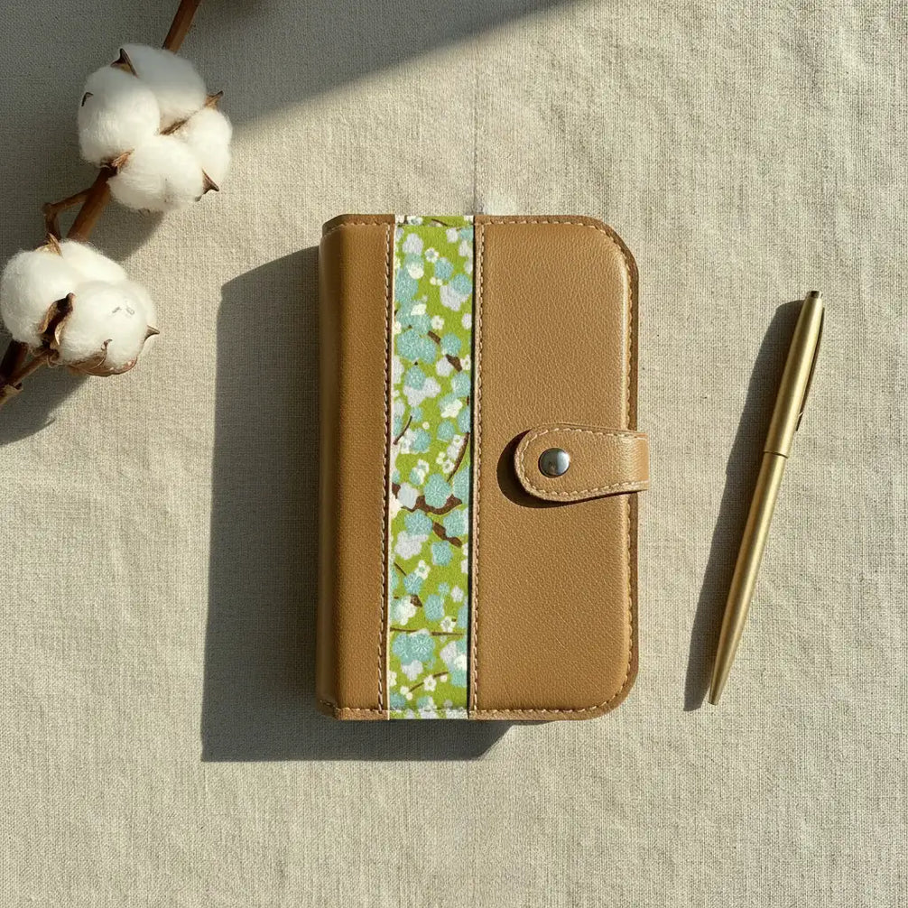 Une prise de vue aérienne élégante d'un classeur modulaire marron doré fermé, disposé au centre. Il est déposé sur la gauche par une branche de capsules de coton naturel et sur la droite par un stylo en métal doré mat. L'ensemble est baigné d'une lumière douce, projetant des ombres, sur un tissu de lin texturé beige.