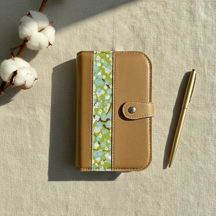 Une prise de vue aérienne élégante d'un classeur modulaire marron doré fermé, disposé au centre. Il est déposé sur la gauche par une branche de capsules de coton naturel et sur la droite par un stylo en métal doré mat. L'ensemble est baigné d'une lumière douce, projetant des ombres, sur un tissu de lin texturé beige.