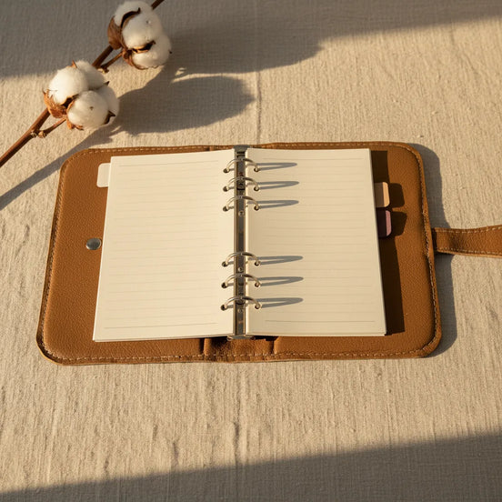 Une vue de l'intérieur d'un classeur modulaire marron doré ouvert, posé sur une nappe en lin texturé. Le mécanisme à 6 anneaux métalliques centraux est visible, tenant des pages de papier ligné de couleur crème. Sur le côté gauche, une poche à soufflet contient des onglets d'indexation colorés. L'ambiance est chaleureuse et organisée.