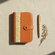Vue de dessus (flat lay) de l'organisateur modulaire A6 en cuir de chèvre orange brûlé et papier japonais, posé sur une toile de lin naturelle à côté d'un stylo doré et d'une branche séchée sous une douce lumière ombragée.