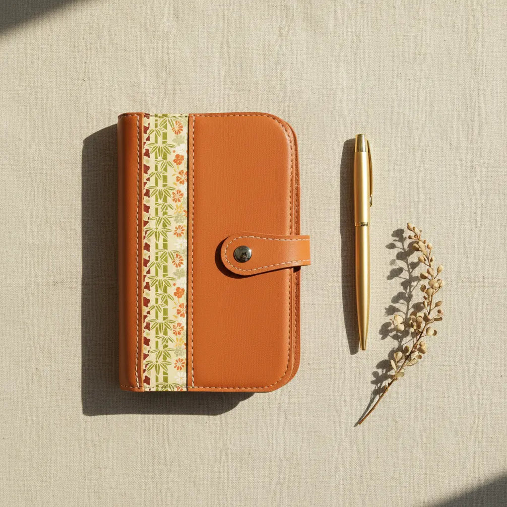 Vue de dessus (flat lay) de l'organisateur modulaire A6 en cuir de chèvre orange brûlé et papier japonais, posé sur une toile de lin naturelle à côté d'un stylo doré et d'une branche séchée sous une douce lumière ombragée.