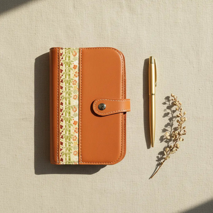 Vue de dessus (flat lay) de l'organisateur modulaire A6 en cuir de chèvre orange brûlé et papier japonais, posé sur une toile de lin naturelle à côté d'un stylo doré et d'une branche séchée sous une douce lumière ombragée.