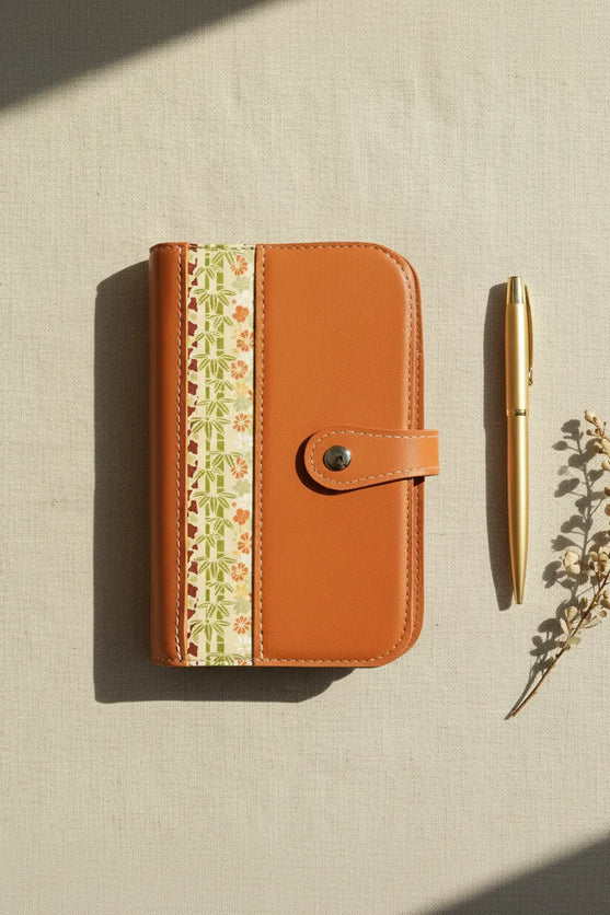 Vue de dessus (flat lay) de l'organisateur modulaire A6 en cuir de chèvre orange brûlé et papier japonais, posé sur une toile de lin naturelle à côté d'un stylo doré et d'une branche séchée sous une douce lumière ombragée.