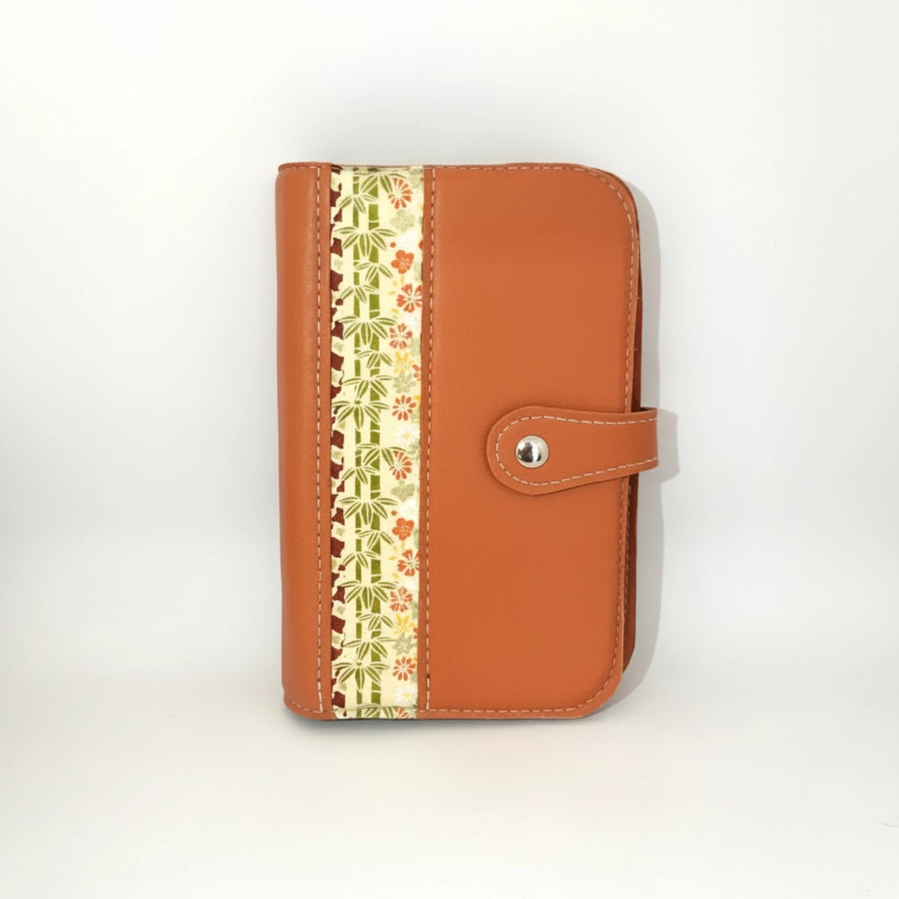 Organisateur en cuir de chèvre couleur orange brûlé fait main, présenté fermé, debout. La couverture avant est bordée par une large bande de papier japonais ornée de motifs complexes de bambous verts, de fleurs rouges et beige. Des points de couture blancs, réalisés à la main à l'alène, soulignent les contours du cuir et fixent le papier. Attache de fermeture en cuir assorti est maintenue par un bouton-aimant en métal argenté brillant.