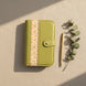 Vue aérienne d'un organisateur modulaire vert pomme fermé, disposé verticalement sur un tissu de fond beige texturé. À sa droite, un stylo fin en métal doré mat et une délicate branche d'eucalyptus sont soigneusement alignés. Une lumière naturelle rasante traverse la scène, créant des ombres douces et une atmosphère sereine.