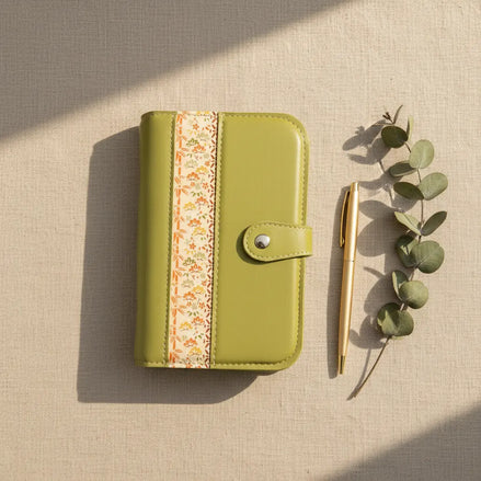 Vue aérienne d'un organisateur modulaire vert pomme fermé, disposé verticalement sur un tissu de fond beige texturé. À sa droite, un stylo fin en métal doré mat et une délicate branche d'eucalyptus sont soigneusement alignés. Une lumière naturelle rasante traverse la scène, créant des ombres douces et une atmosphère sereine.
