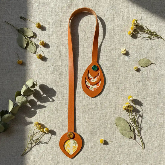 Marque-page artisanal en cuir cognac posé à plat en forme de U sur fond lin naturel, ses deux extrémités à médaillons ovales découpés révélant un papier japonais à motif floral orange et crème, ornés de rivets vert émeraude et ambré, entouré de fleurs séchées jaunes et de feuilles d'eucalyptus en lumière matinale rasante.
