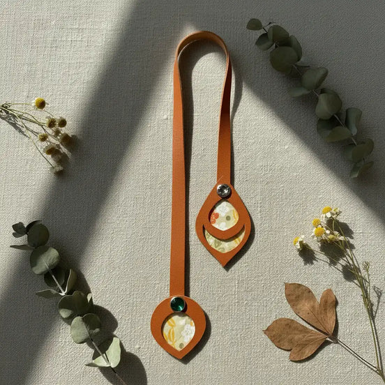 Flat lay d'un marque-page en cuir cognac déployé sur fond lin, présentant ses deux extrémités à médaillons découpés avec incrustations de tissu japonais fleuri jaune-orange, rivets cristal et vert, entouré de fleurs séchées et feuilles d'eucalyptus.