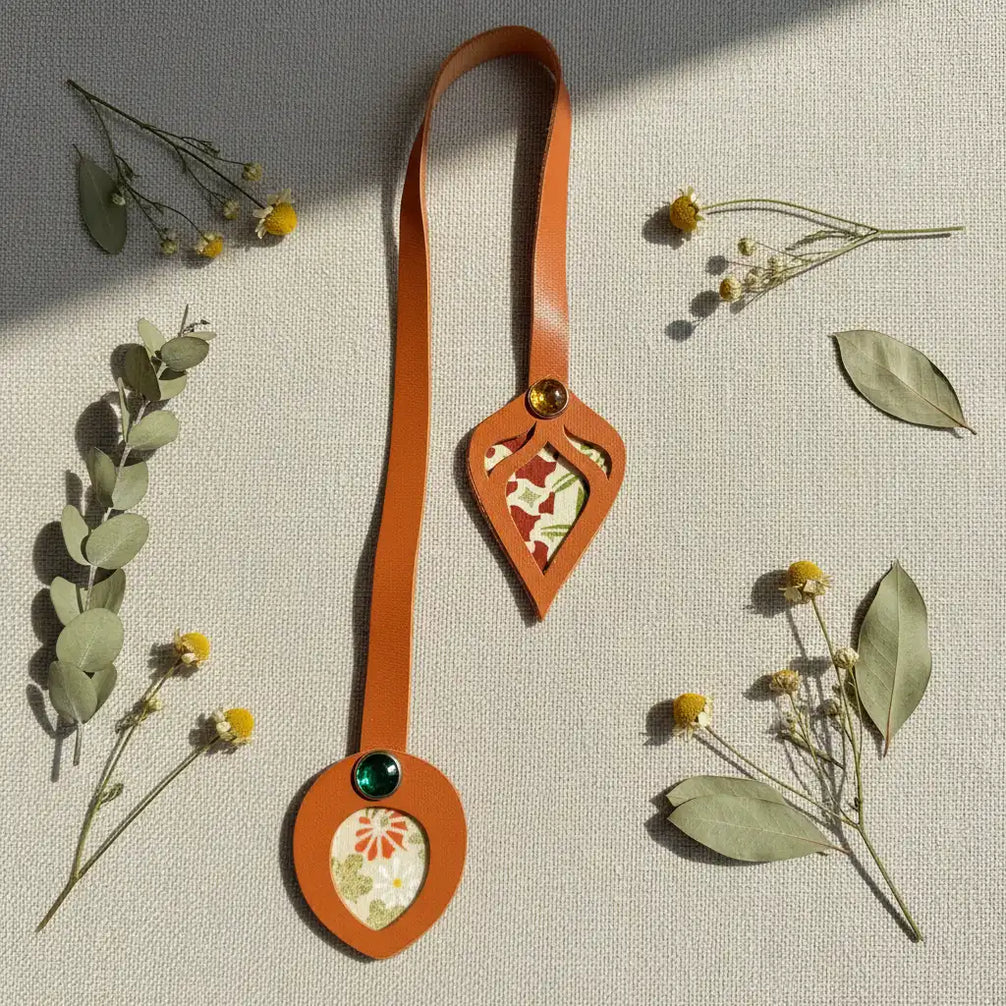 Marque-page en cuir cognac à découpe feuille et médaillon, ornés de tissus japonais fleuris et de cabochons colorés, entourés de fleurs séchées et feuilles d'eucalyptus sur fond lin naturel.
