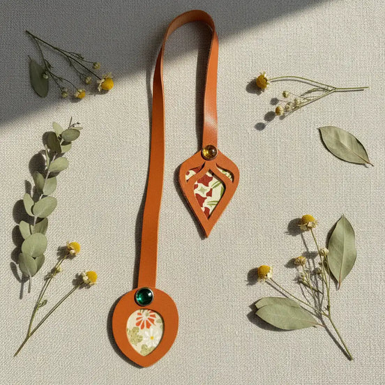Marque-page en cuir cognac à découpe feuille et médaillon, ornés de tissus japonais fleuris et de cabochons colorés, entourés de fleurs séchées et feuilles d'eucalyptus sur fond lin naturel.
